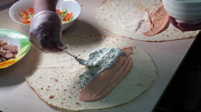 Gloved hand adding white garlic herb sauce next to pink sauce on a flatbread. Assembling a homemade shawarma or wrap with meat and vegetables.