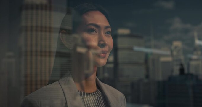Asian Businesswoman Stands by a High-rise Window, Looking at Skyscrapers and Clouds Beyond the Glass as City. Ambition, Confidence, Modern Success. Sunny Cinematic Reflection Portrait