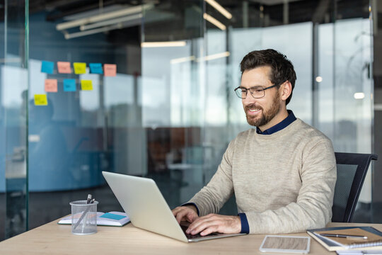 Young professional man in glasses smiling while focused on his laptop at a modern office desk, conveying productivity, confidence and a comfortable, smart-casual work environment