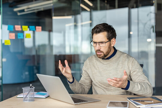 Stressed businessman in glasses sitting at a modern office desk, frustrated and gesturing toward a malfunctioning laptop while struggling with deadline pressure and tech failure