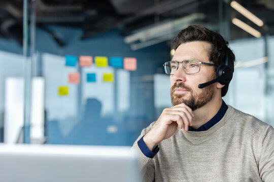 Bearded call center agent with glasses and headset in a modern office, looking away thoughtfully while providing customer support and technical assistance with concentration and professionalism