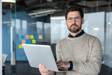 Fototapeta premium Professional man with a beard and glasses using a laptop, standing in a modern office environment, representing business, technology, and corporate success