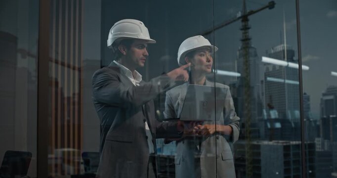 Caucasian Male and Asian Female Construction Engineers in Hard Hats Review a Laptop by Window, Focused on Project Finance and Growth. Reflections Reveal Big City Real Estate Buildings and Cranes. 