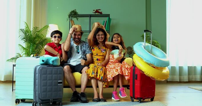 Cheerful Indian family of four in tourist attire sitting on a sofa with suitcases. Throwing caps in the air in happiness before their summer vacation trip