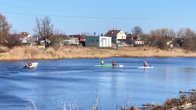 people kayaking on river in early spring with supervisor in boat, village houses and trees on opposite bank