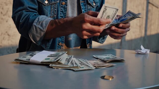 Counting cash and stacking dollar bills. Person counts money with hand over table. Pile of cash sits next to stack. Jacket sleeve visible during transaction. Concept of finance exchange and income.