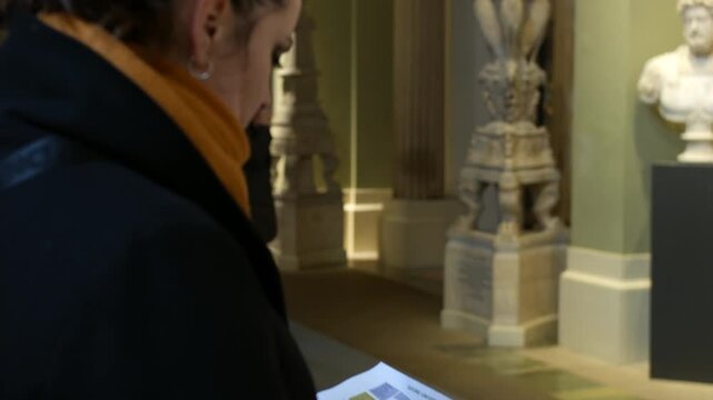 Woman holding a museum map while walking through a sculpture gallery lined with classical statues on pedestals. Interior museum corridor with visitors exploring exhibits and reading the layout guide