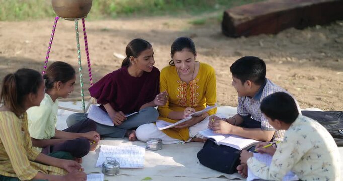 Indian villager girl boy sitting on mat do study read book outdoor rural area ground place. Happy group child hold pencil writing notes talk gossip enjoy desi life new day outside open class field