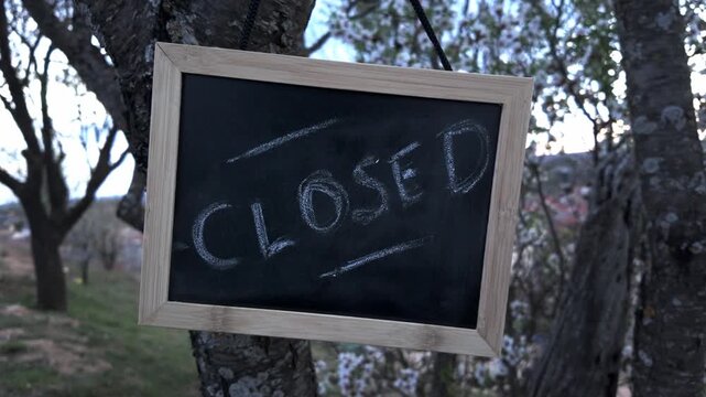 Chalkboard sign reading CLOSED hanging from a tree branch. Concept of business closure for rest, health leave, holidays, or temporary pause in activity.