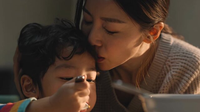 Parent kissing a child on the forehead while the child eats, expressing love, care, and nurturing family moment during mealtime.