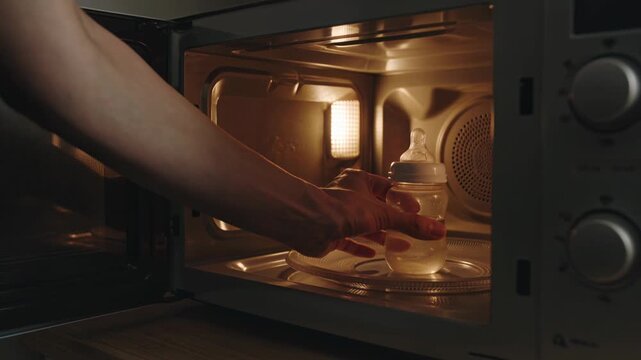Hand placing a baby bottle inside a microwave for warming, illustrating childcare routine and preparation of infant feeding indoors.