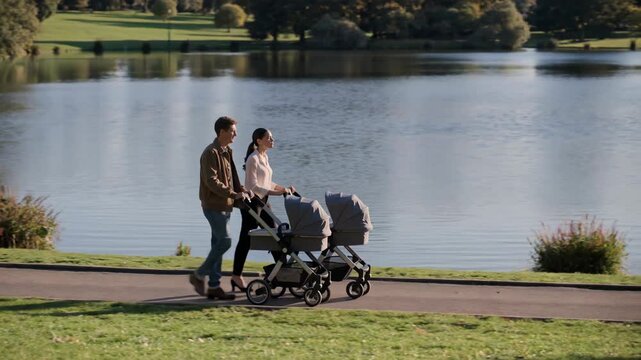 Couple walking with strollers along lakeside path in green park, enjoying peaceful family outing and calm outdoor environment.