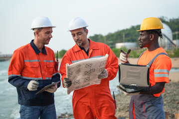 Engineering team in safety gear reviewing technical blueprints, using a tablet, and rugged laptop...