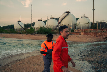 Obraz premium Diverse professional engineers in safety gear walking at a coastal oil and gas refinery plant with large spherical storage tanks. Industrial energy sector workspace and technical teamwork