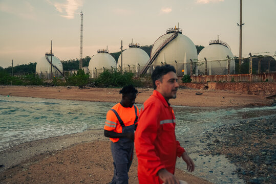Diverse professional engineers in safety gear walking at a coastal oil and gas refinery plant with large spherical storage tanks. Industrial energy sector workspace and technical teamwork