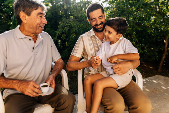 Grandfather, adult son and young grandson sitting outdoors and interacting with affection, representing family bonding and multigenerational connection.