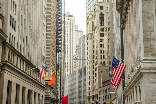 Historic financial buildings and skyscrapers line Wall Street in New York City financial district. Multiple American flags hang along the street representing business, finance, and the United States