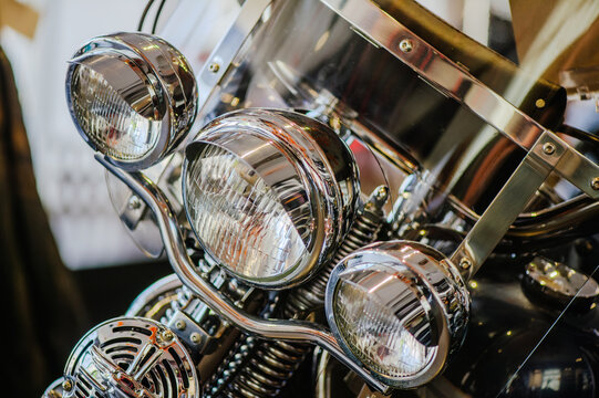 A horizontal close-up shot of the front lighting assembly of a motorcycle, featuring three round chrome headlights and a transparent windshield with metallic supports.