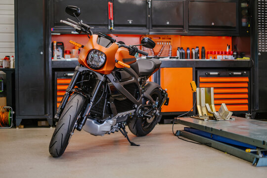 A wide horizontal shot of a sleek orange electric motorcycle parked in a clean, organized garage with black and orange tool cabinets and a hydraulic lift.