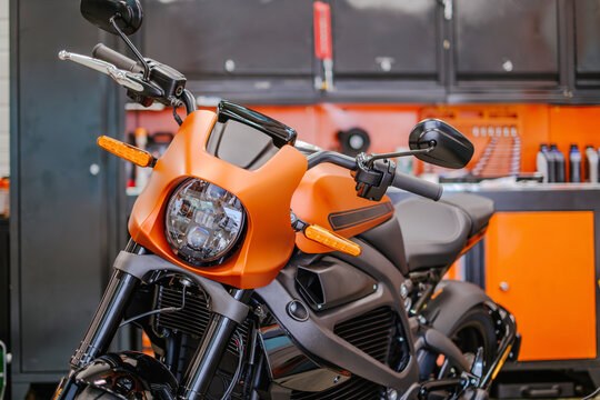 A medium horizontal shot focusing on the front fairing,  headlight, and  matte orange electric motorcycle. Background shows a professional garage setting with orange and black tool storage.