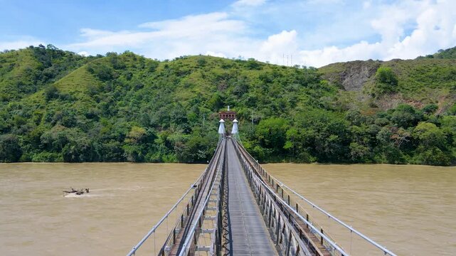 Long wooden suspension bridge crossing a muddy brown river toward a small tower, framed by green hills and blue sky with scattered clouds.