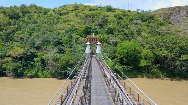 Drone flies along a narrow suspension bridge toward two white conical towers and a red brick gateway over a brown river, framed by lush green hills under blue sky.
