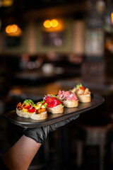 Fototapeta premium A gloved hand holds a black platter with four assorted gourmet bruschettas. The toppings include tomatoes, tuna, and roast beef, set against a blurry dark restaurant background