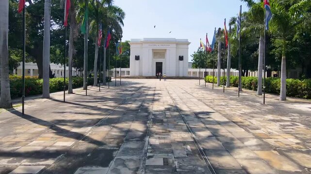 A white mausoleum reading 'Colombia al Libertador' at the end of a palm-lined paved plaza with rows of flagpoles and visitors on the steps under a clear blue sky.