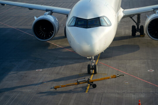 White passenger airplane parked on a gray airport tarmac. A yellow tow bar is positioned near the front landing gear, ready for ground operations