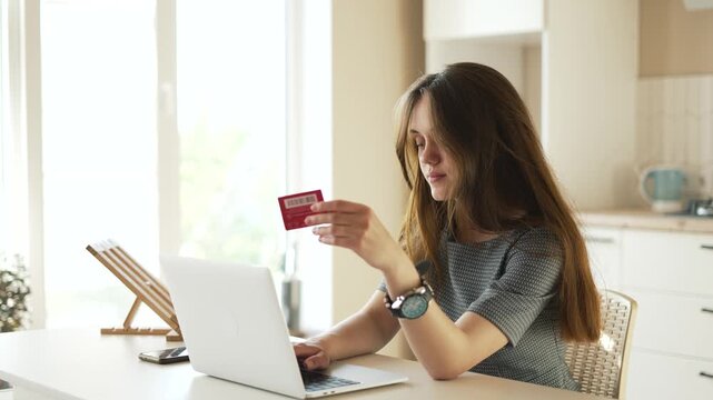 Woman holding credit card using laptop, shopping online at kitchen table in home, checking payment and purchase details on internet store while planning delivery and comparing price and order.