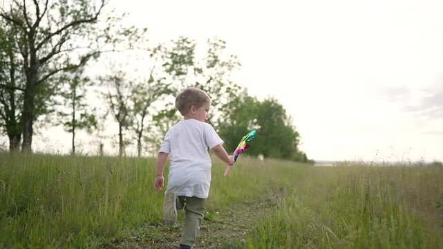 boy running through field grass holding colorful pinwheel toy child back view in tall grass near tree line under bright sky summer nature outdoor play carefree movement summer play pinwheel child