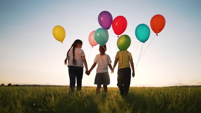 Children hold balloon and walk through open grass field toward sunset while colorful balloon cluster floats in sky creating silhouette of child and friend in small group bathed in warm light