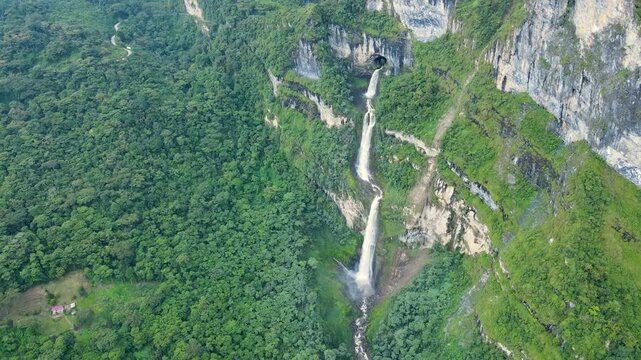 Drone shot of multi-tiered Tisquizoque waterfall pouring from a cave over limestone cliffs into a green forest valley; small red-roof house visible in a clearing.