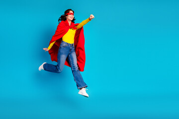 Obraz premium Young woman superhero leaps in bright red cape against blue studio background expressing joy and confidence