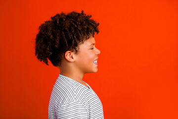 Smiling boy profile portrait with curly hair against a vibrant orange background, cheerful child looking right and laughing in a casual striped shirt studio headshot © deagreez
