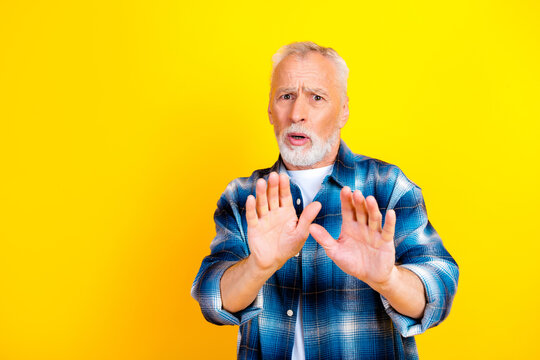 Senior man in blue plaid shirt holding hands up in stop gesture with surprised expression against bright yellow background conveying caution and refusal