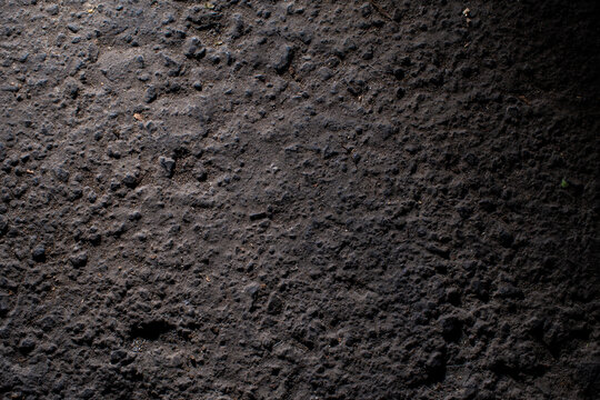 Detailed shot of dark earth ground or asphalt showcasing gritty texture and uneven surface under high contrast side lighting for a dramatic effect.
