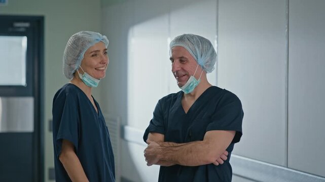 Medium shot of Caucasian nurses in deep blue scrubs having small talk and joking while standing in hospital hall