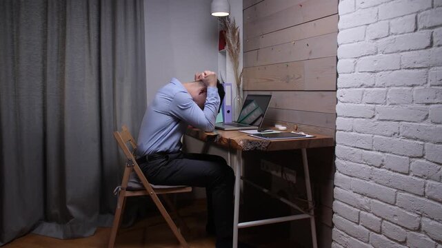 Man sits at a rustic wooden desk in a home office, holding his head in his hands, overwhelmed by work pressure, exhaustion and emotional stress while staring at a laptop