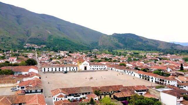 Aerial drone sweep of Villa de Leyva's vast cobblestone main square, white church with yellow trim and red tile roofs set against green mountains.