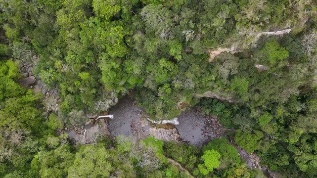 Overhead drone captures two small waterfalls spilling into rock pools beneath a dense green canopy and a winding stream in Villeta, Cundinamarca.