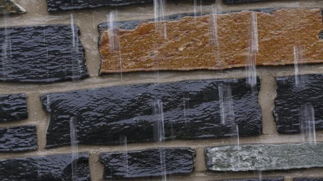 Raindrops fall vertically onto a brick wall. Bricks show black, brown, and gray tones. Water streaks down the textured surface. Close-up view captures rain's impact