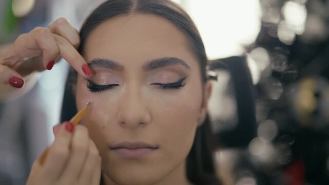 A makeup artist applies false eyelashes to a young woman's eye. The client enjoys an enhancing beauty treatment at the salon.