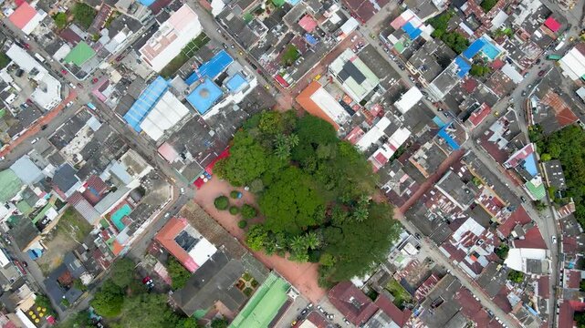 Drone overhead of a square plaza dense with green trees and palms, surrounded by colorful rooftops and intersecting streets in Villeta, Cundinamarca