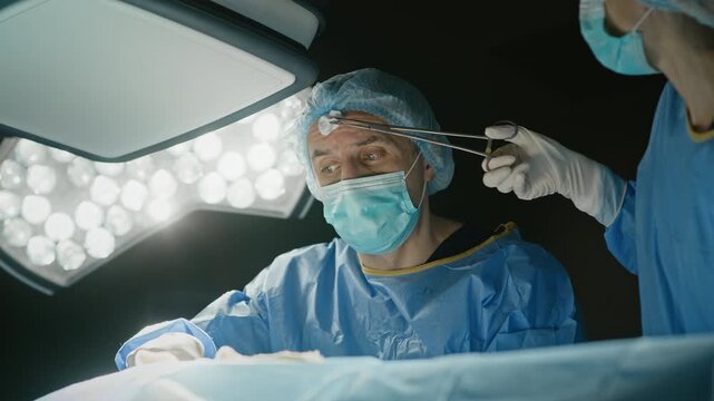 Low angle shot of assistant dabbing sweat from forehead of surgeon who performing surgery on patient
