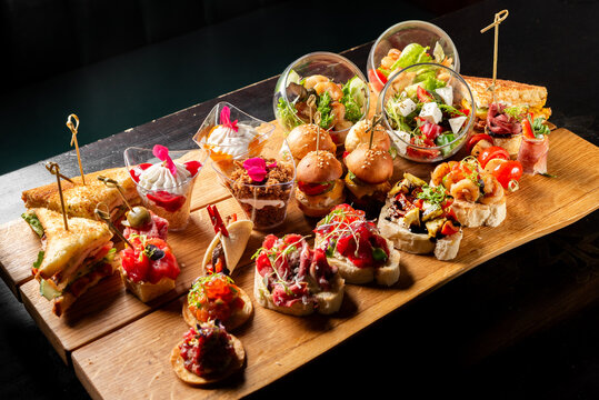 Assorted catering snacks and appetizers on a wooden board: bruschettas, mini burgers, sandwiches, salads, and desserts in glasses. Elegant finger food for party or event
