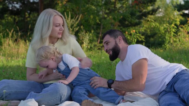 Sunny picnic with white family cuddling toddler on blanket, mother smiling, father reaching playfully, golden light, meadow backdrop, casual jeans and yellow top, affectionate interaction,