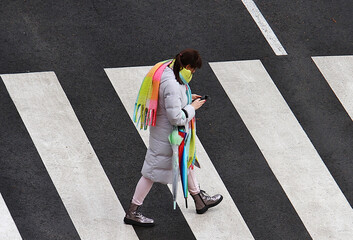 Mujer con bufanda de colores, paraguas y abrigo cruzando por paso de peatones y mirando teléfono móvil © Diego