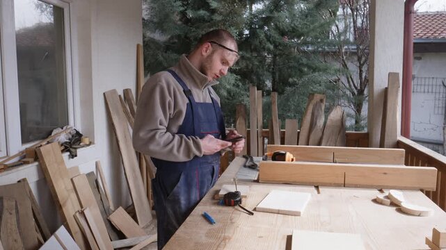 A male woodworker checks his smartphone while taking precise measurements of wooden pieces. He carefully plans his next steps in a dedicated home workshop environment.