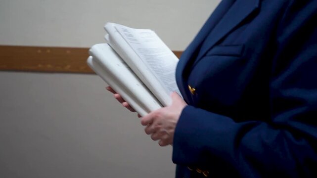 Close-up shot of a woman's hands in a blue uniform jacket holding a thick stack of white papers. The woman is standing in a hallway. Focus on the texture of the paper and the details of the official u